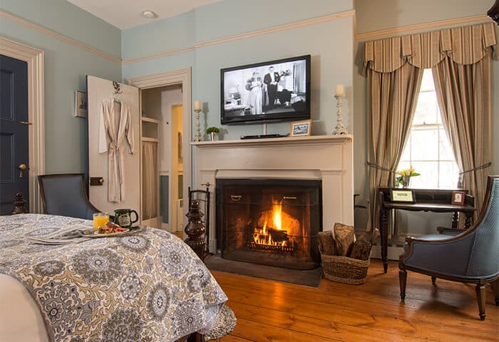 Guestroom features a neatly made bed in front of fireplace and television; walls are a soft blue with hardwood flooring