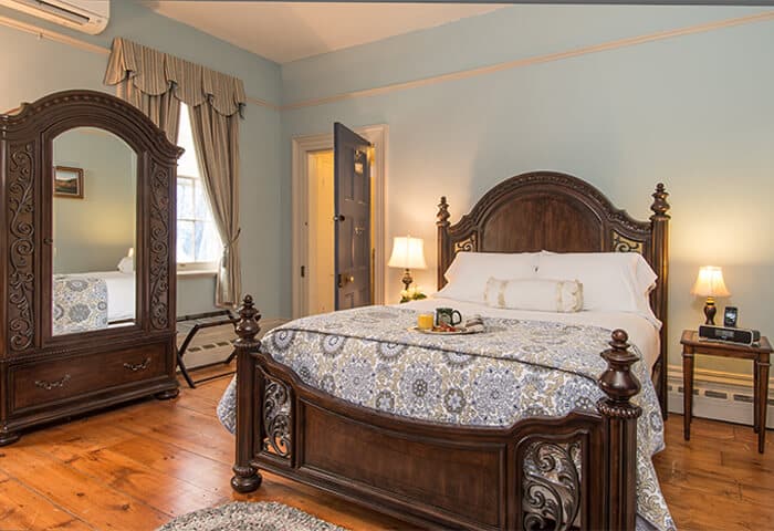 Guestroom features luxurious bedding against dark wood bedframe; walls are a soft blue with hardwood flooring