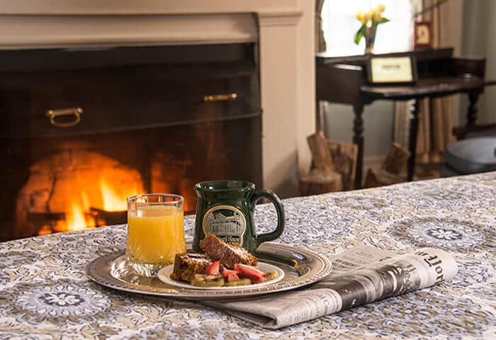 A silver tray holds coffee, orange juice, and fruit on a neatly made bed; a roaring fire is in background