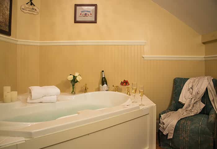 Bathroom features luxurious white tub with cream colored walls; small chair is to the side with spa amenities at hand