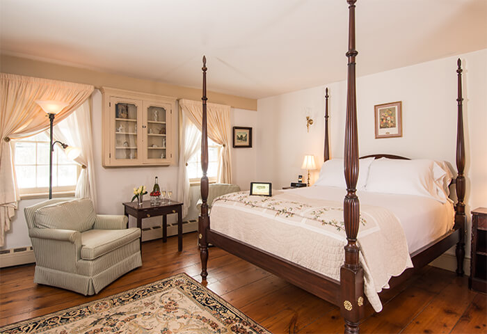 Charming bedroom with cream walls features inviting four post bed with white linens and quilt; comfy chair to the side