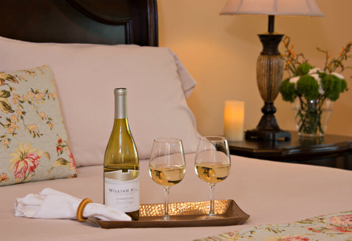 A neatly made queen bed with bright white linens features a tray with white wine and wine glasses