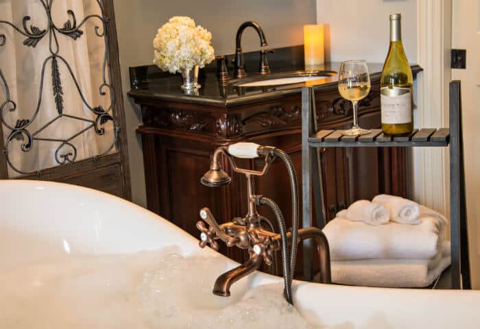 Sage blue bathroom with dark wooden vanity and white antique bathtub with towel stand featuring delicious wine