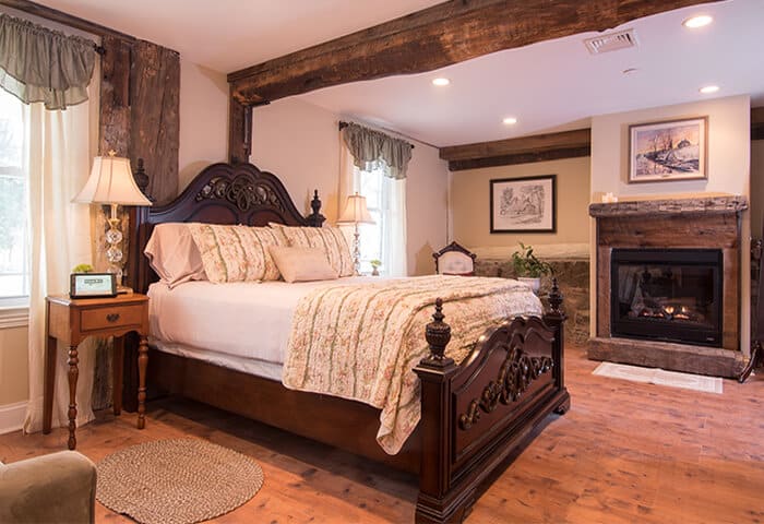 Inviting guestroom features rustic fireplace with elegant bedding on a dark wood bedframe; cheery light flows in from windows