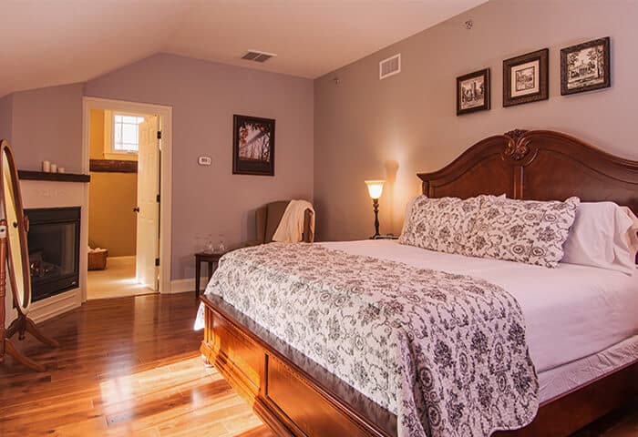 Inviting guestroom features corner fireplace and hardwood floors and neatly made bed. Wall color is a pleasing velvety heather