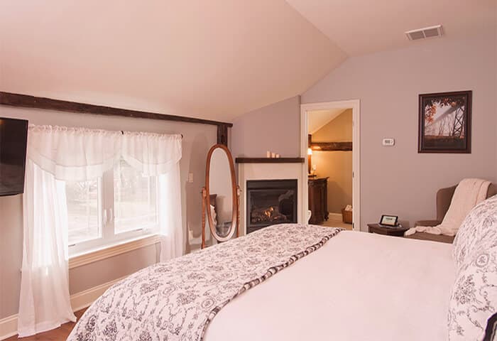 Inviting guestroom features corner fireplace, bright white window curtains and neatly made bed. Wall color is a pleasing velvety heather