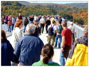 A large crowd of people walking over a long bridge with beautiful fall landscape in the background 