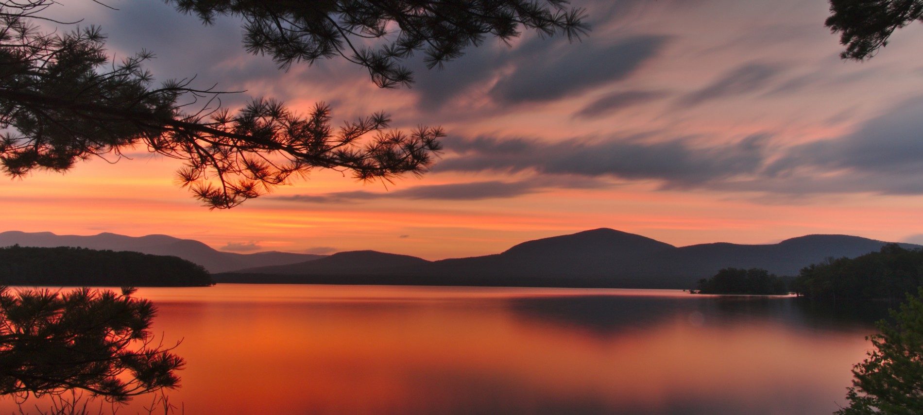 Large reservoir with view of mountain range in background surrounded by bright orange skies at dusk