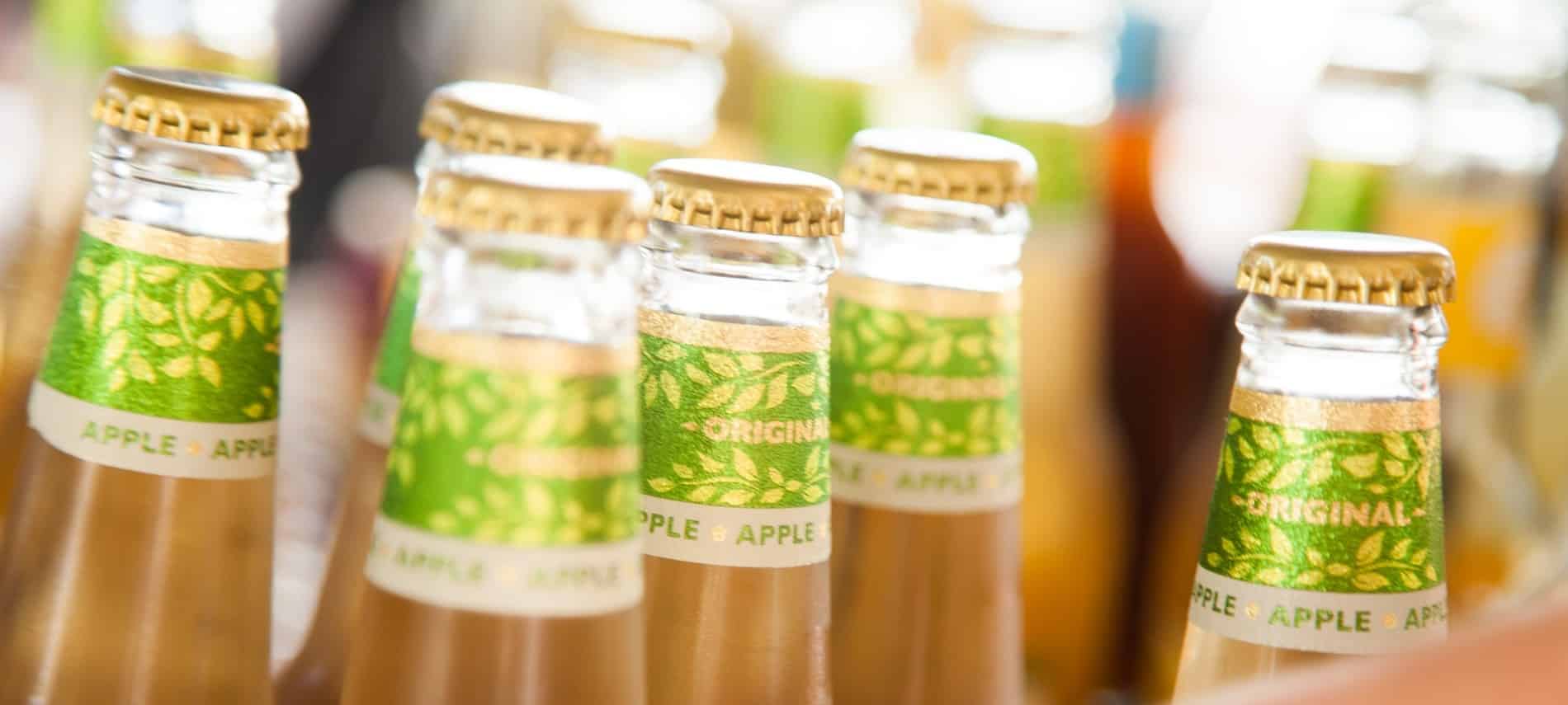 Six bottles with gold caps and light brown beverage with green "apple" labels