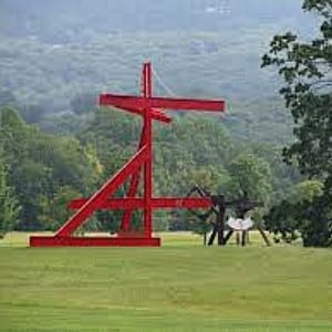 Large red metal art sculpture and small black sculpture sitting in an open field with forest in background