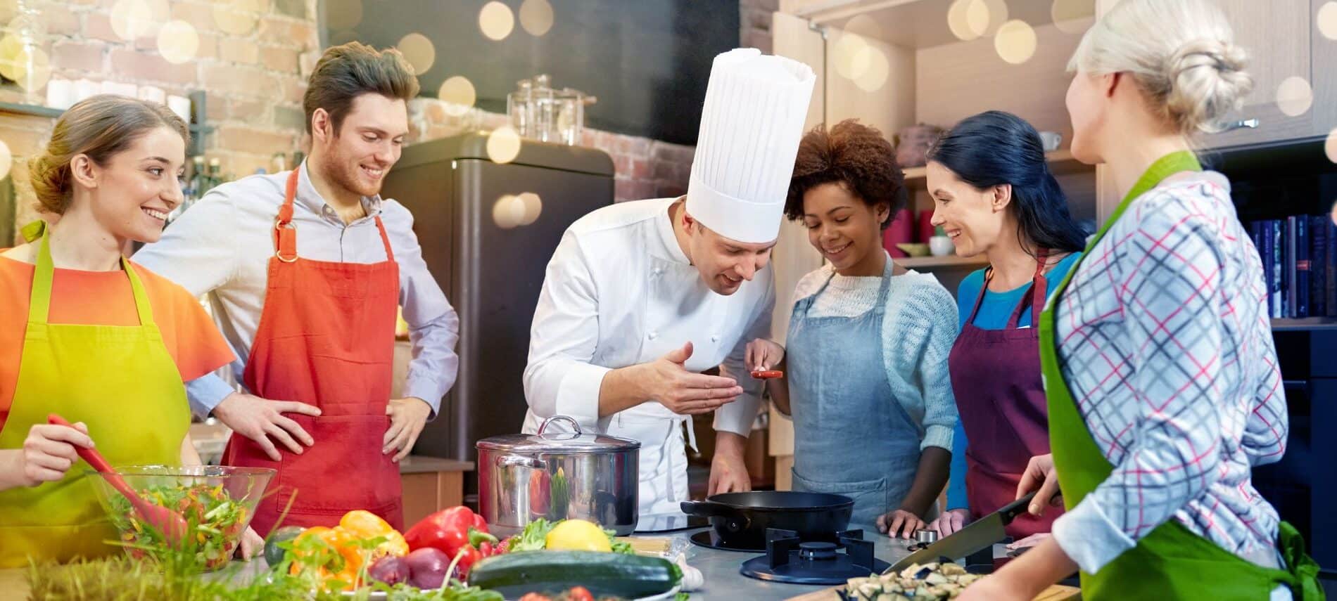 Five people in aprons standing around a commercial kitchen station watching a chef in chef toque explain something on the stove.