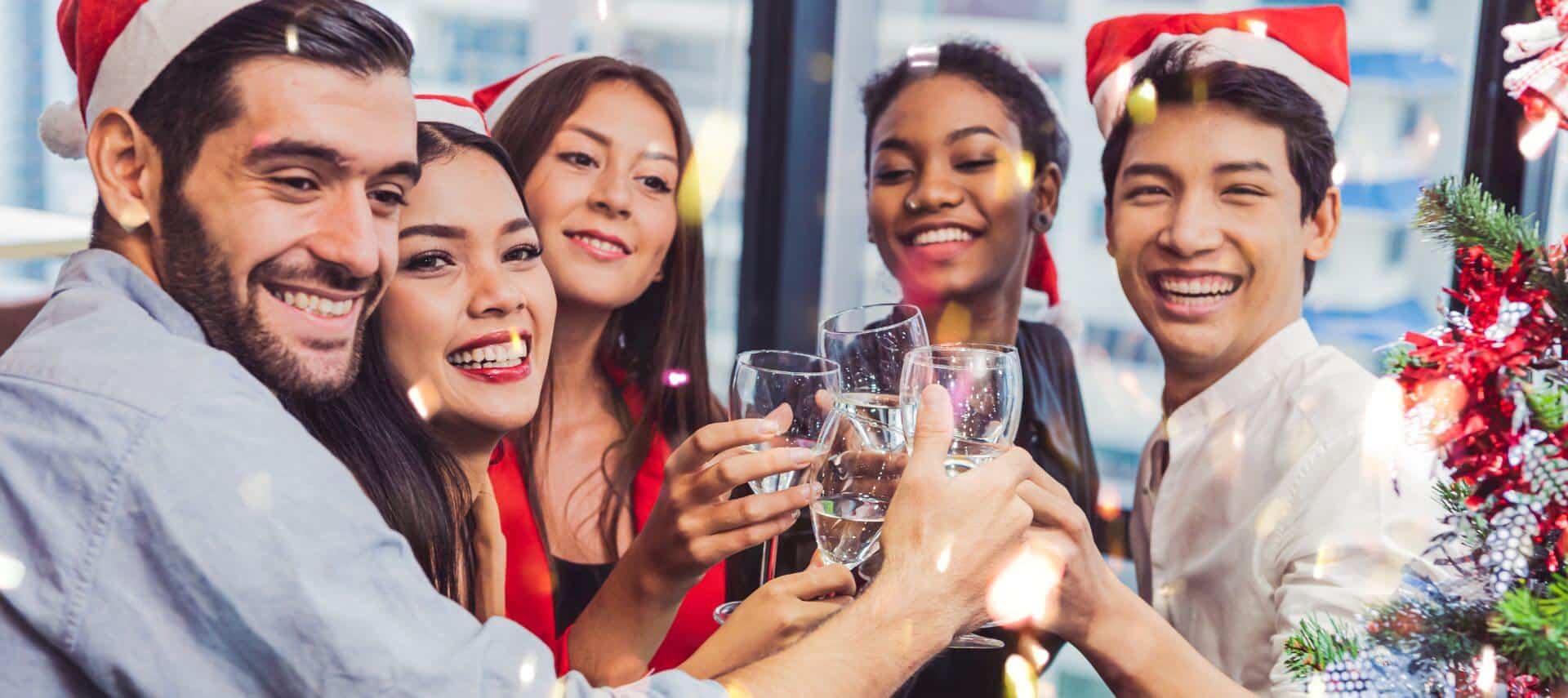 a group of friends wearing red and white santa hats toasting wine next to a christmas tree