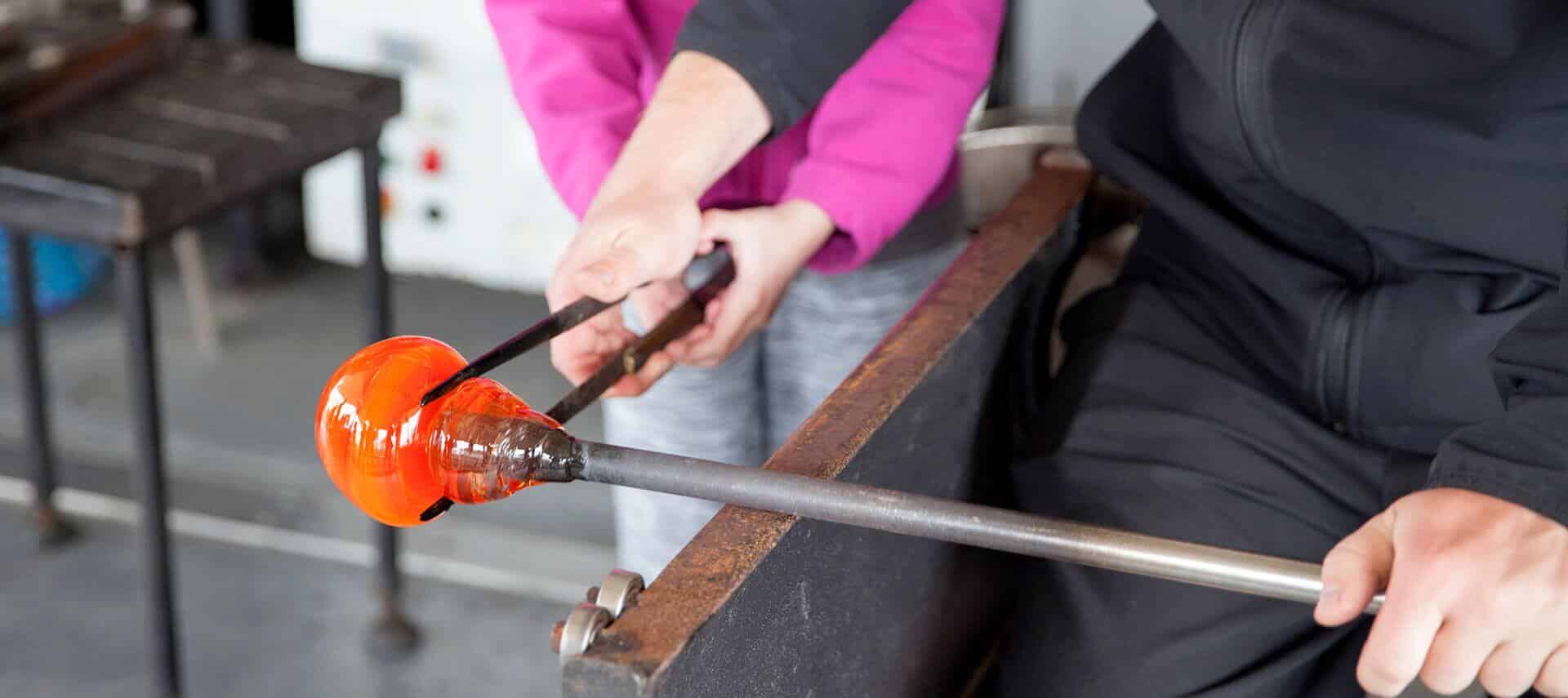 Close-up of a man and a woman in a glass blowing class holding a molten glass project with tongs and rod