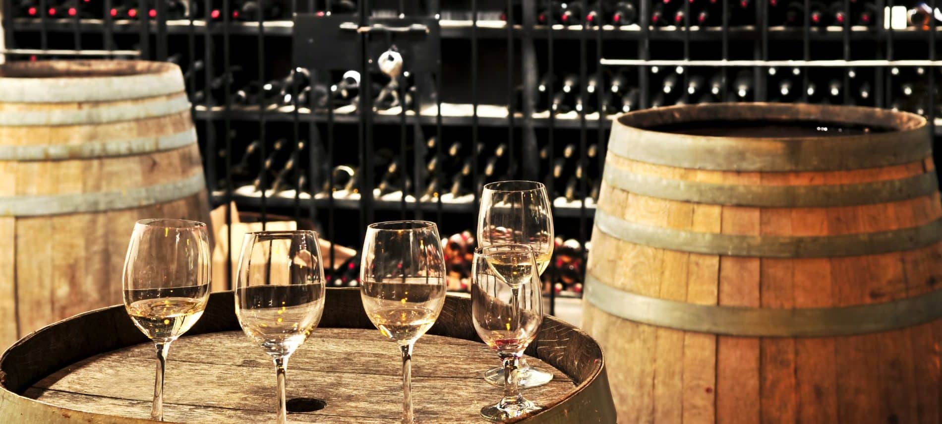 Five partially filled wine glasses on top of an oak wine barrel. There are barrels on either side, with many racks of wine behind.