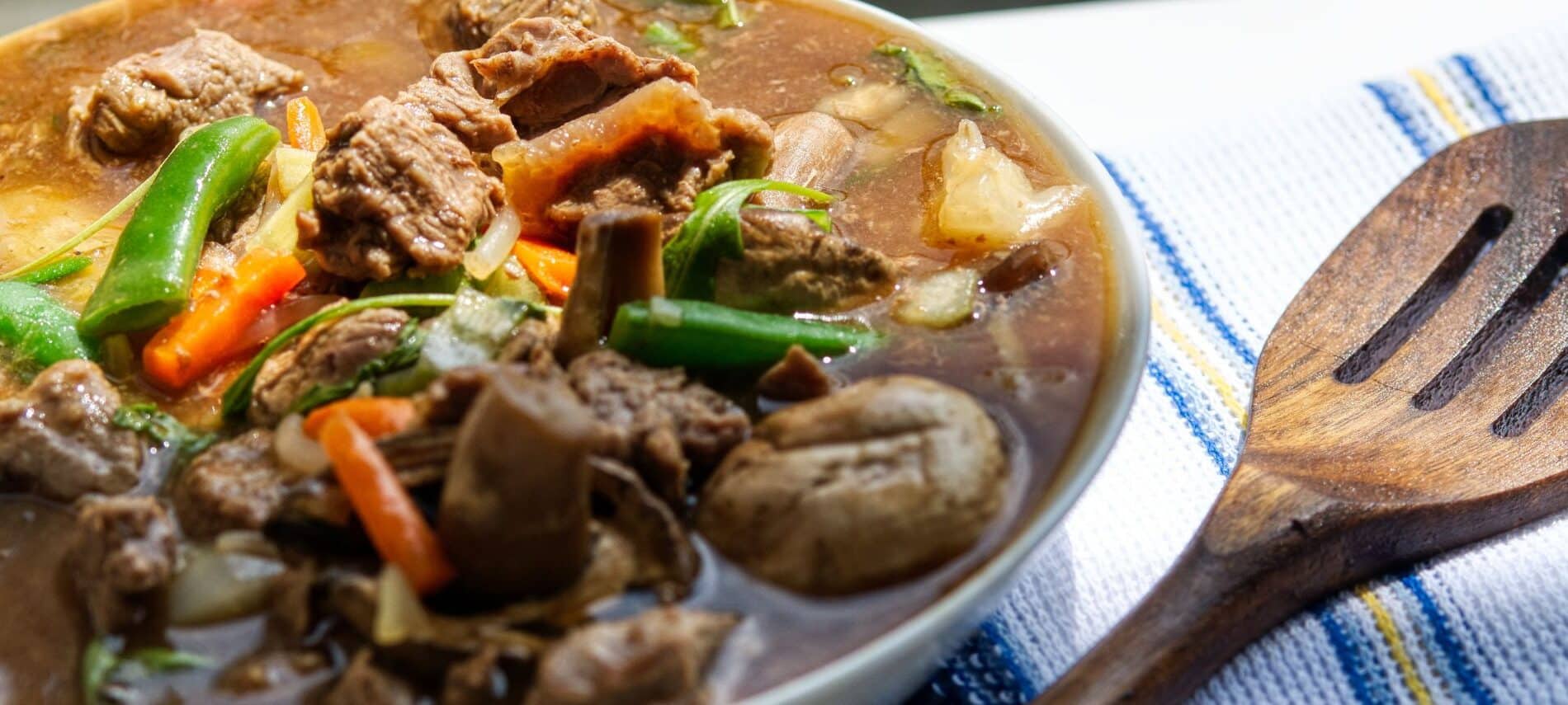 a bowl of irish stew with beef, carrots, green beans, onion, and potatoes with a large slotted wooden spoon next to it on a blue and white tablecloth