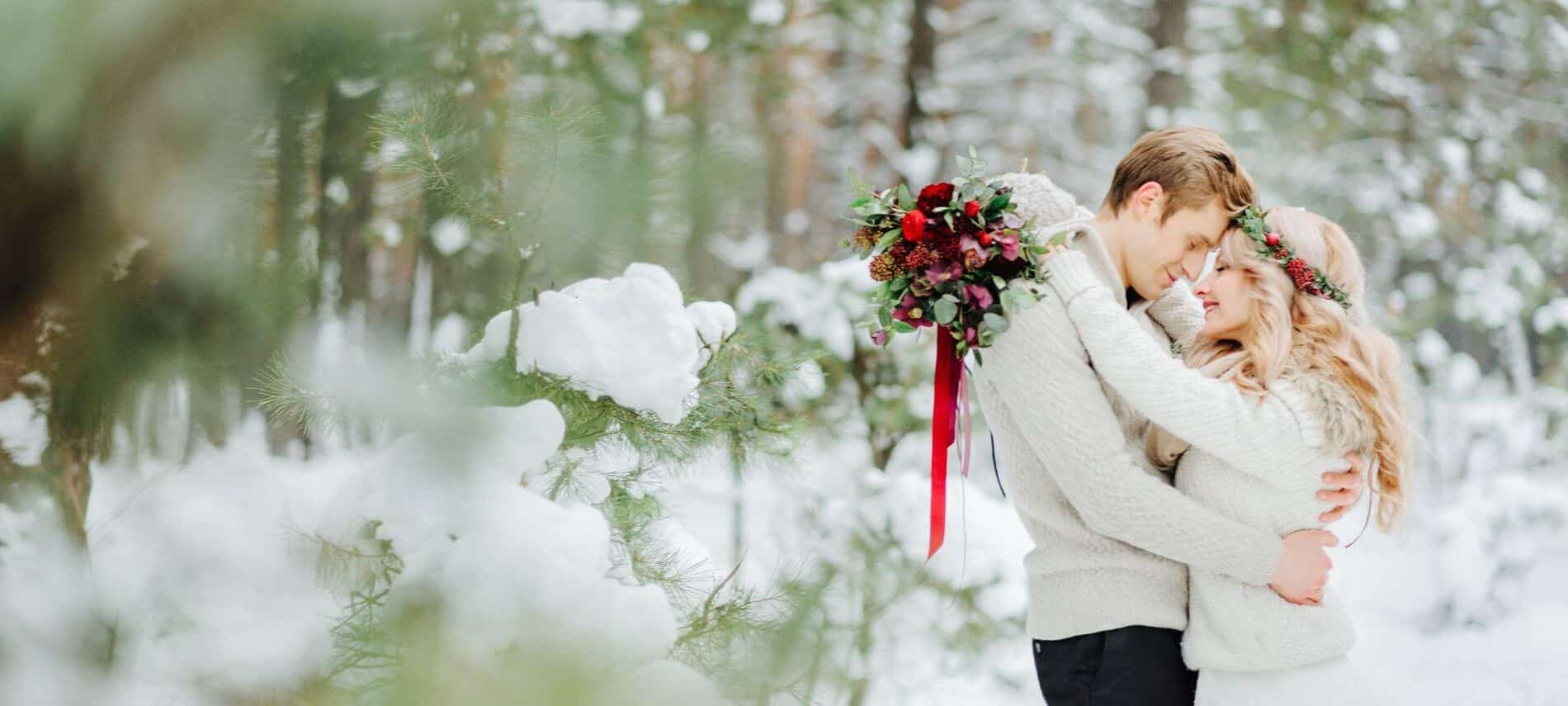 romantic couple standing outside and embracing while woman holds red roses and wears red rose crown, snow-covered trees surround them
