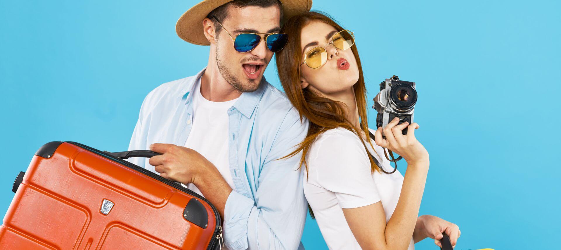 romantic traveling couple standing back-to-back, man holds red suitcase and woman blows kiss while holding camera