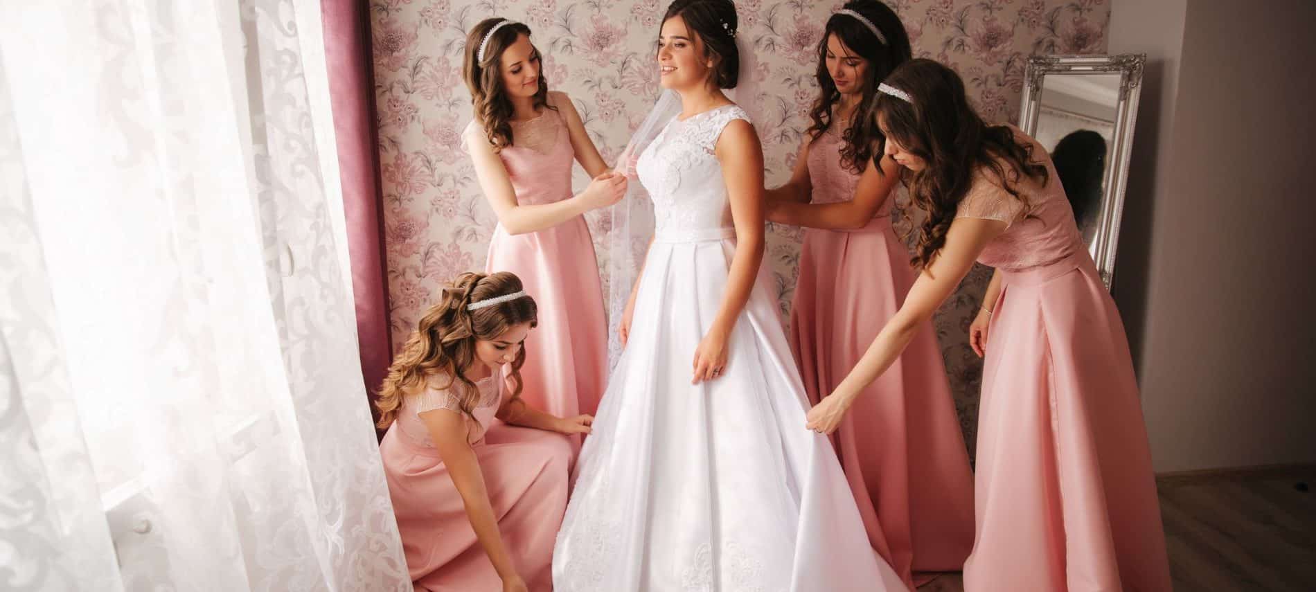 Bridesmaids dressed in pink helping bride finish dressing next to a mirror and open window