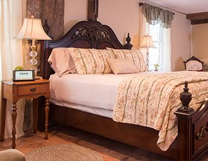 Inviting guestroom features rustic fireplace with elegant bedding on a dark wood bedframe; cheery light flows in from windows