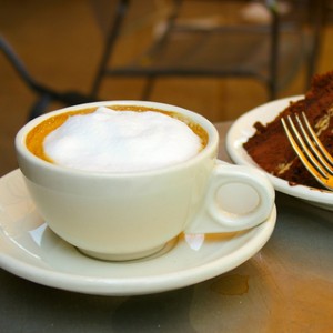 White mug of coffee with whipped foam on top next to plate of chocolate cake