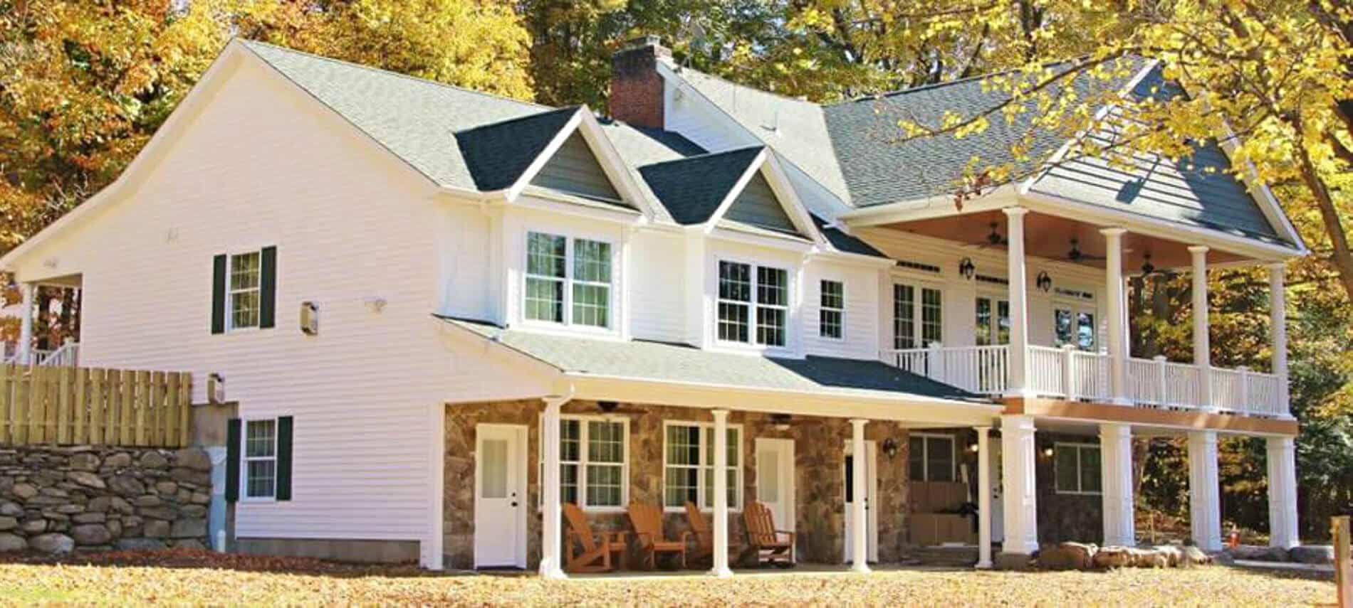 Exterior view of large white house accented with natural stone, among golden trees of autumn; featuring comfy seating on porches