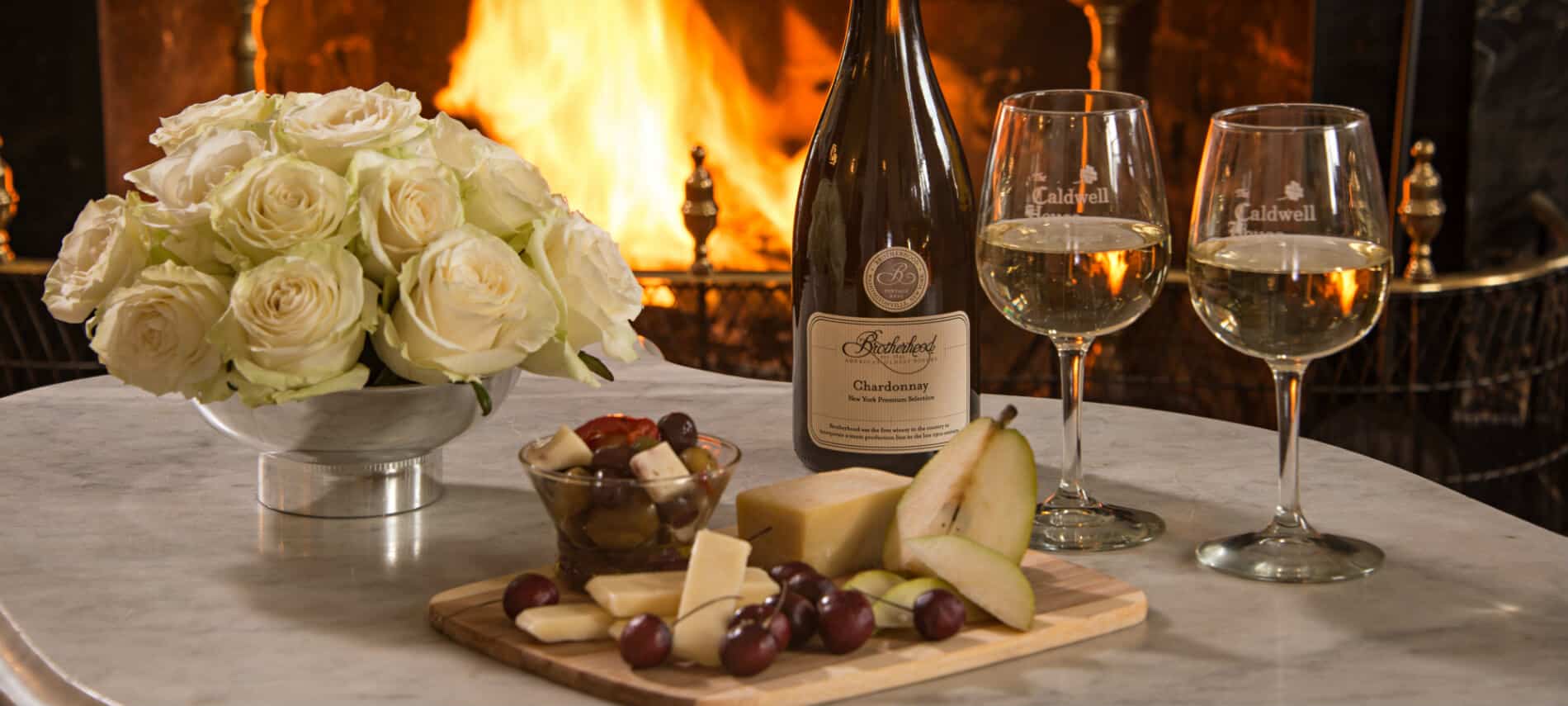 A white marble table displays fruit and cheese on tray, a bottle of Chardonnay and wine glasses; fireplace in background