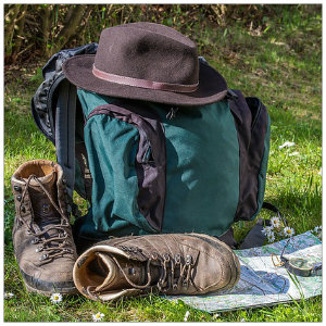 Backpack, hiking shoes, hat, map, and compass sitting on the ground in green wooded area