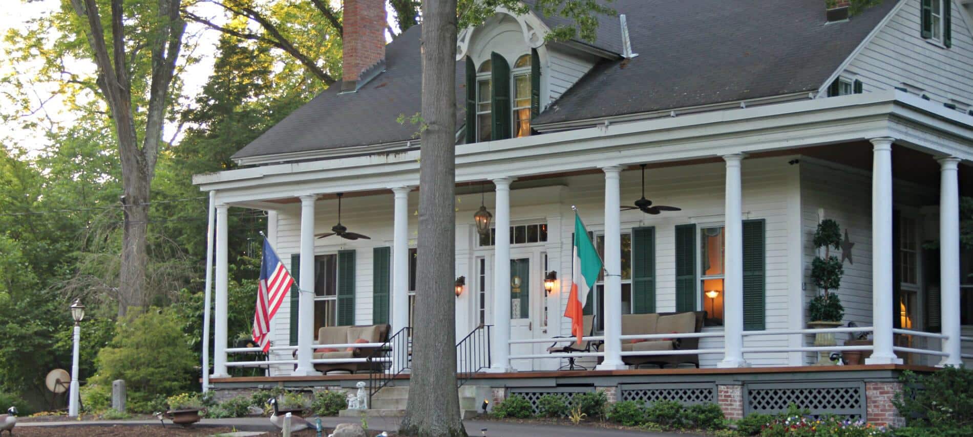 Front view of two-story white house with green shutters surround by woods, porch with furniture, American Flag.