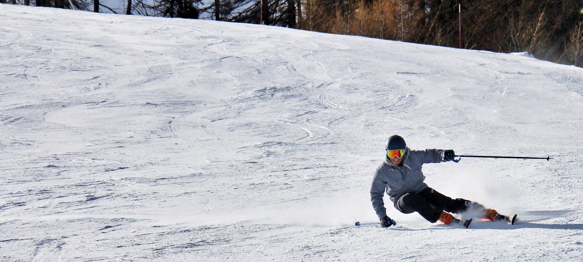 One lone skier in black pants and a blue jacket skiing down a large snow-covered hill with trees behind