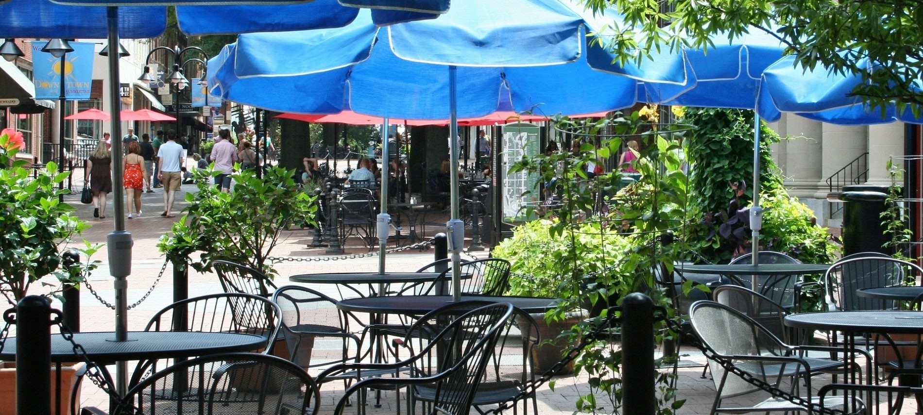 Outdoor dining area with several black tables under blue umbrellas on a street with people walking