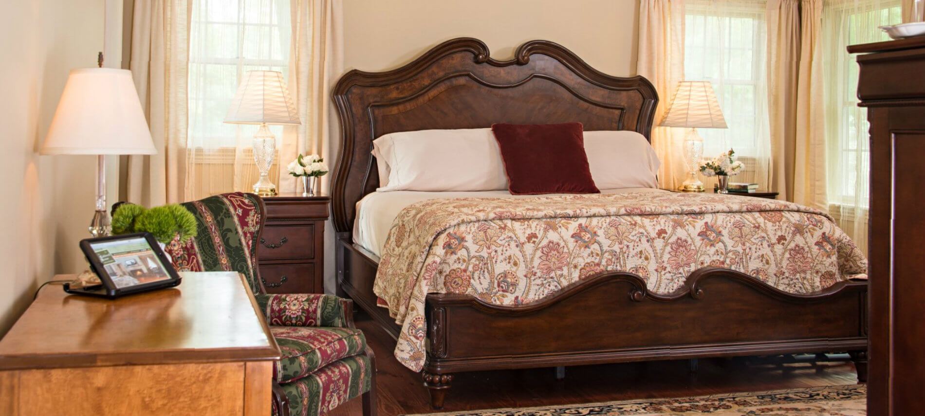 Cozy bedroom with cream colored walls has dark wood furniture; includes inviting bed with quilt and comfortable side chair